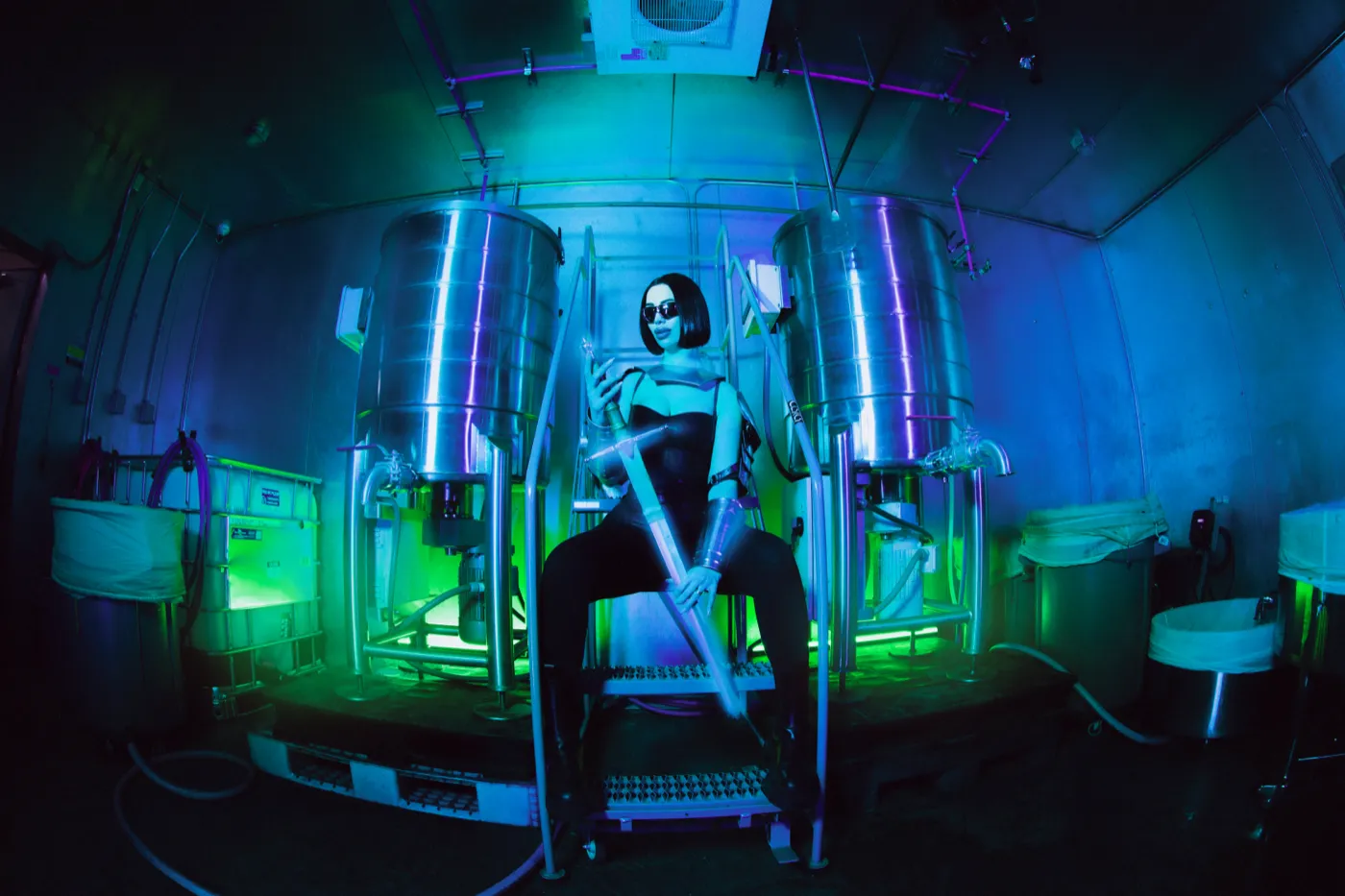 A woman in dark glasses poses between two steel tanks in a blue and green lit rosin lab