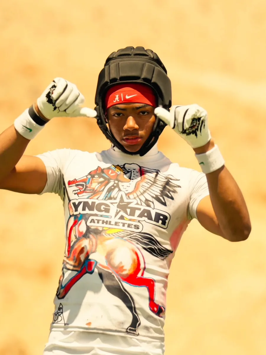 A young athlete in a padded helmet and a YNGSTAR Athletes tee points both thumbs at himself, desert behind