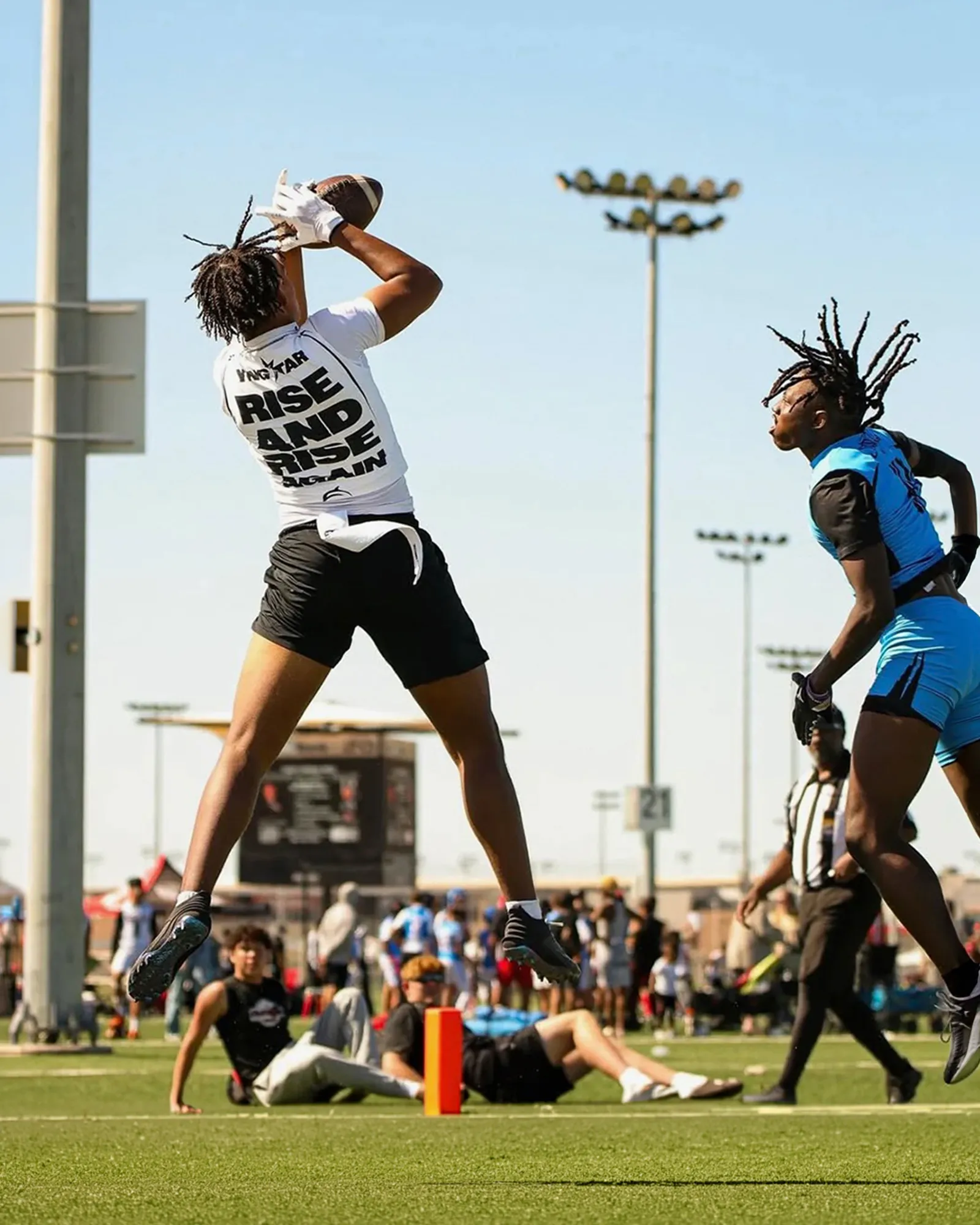 A player in a YNGSTAR "Rise and Rise Again" tee leaps to catch a football on a sunny tournament field